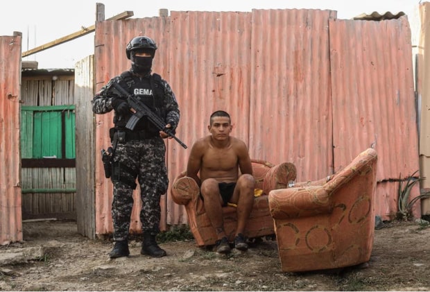 officer with a gun standing next to a man sitting on a couch